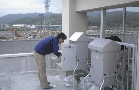 宮城県石巻商業高校屋上での大気粉じんのサンプリング風景と震災がれき置き場（奥）の写真