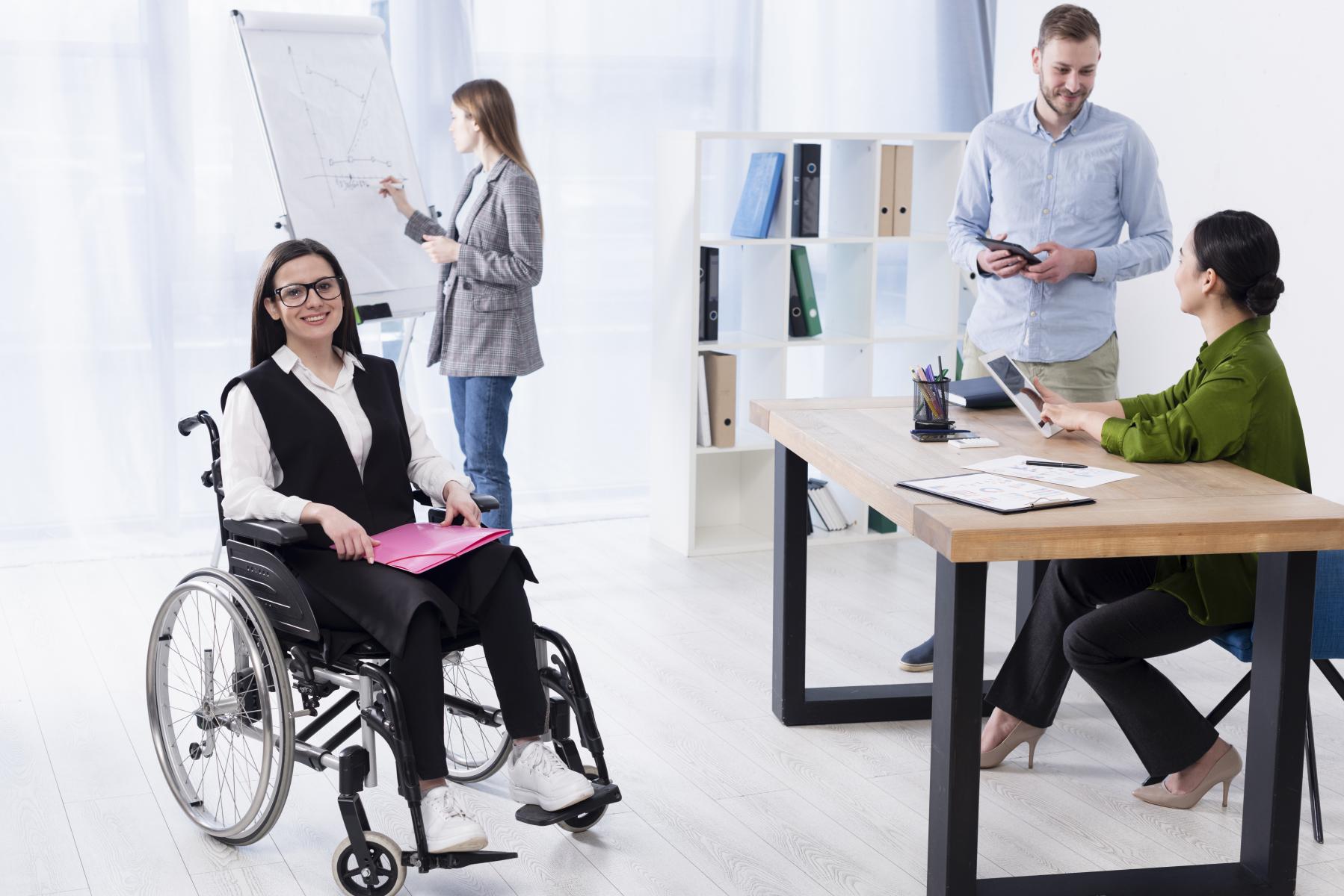 多様な人種で構成されるオフィスワーカーが一緒に仕事をしているオフィスの風景。車椅子に乗っている障がい者の女性がファイルホルダーを膝に乗せ、こちらに微笑んでいる